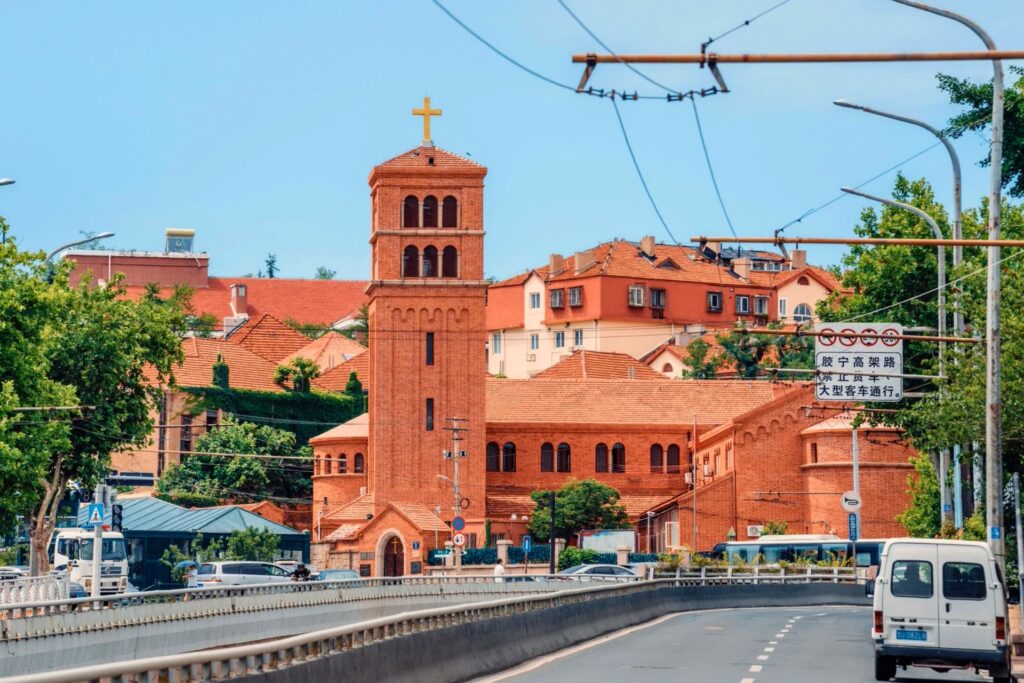 Église en briques rouges dans la vieille ville de Qingdao, entourée de bâtiments aux toits rouges et de verdure, avec une route au premier plan