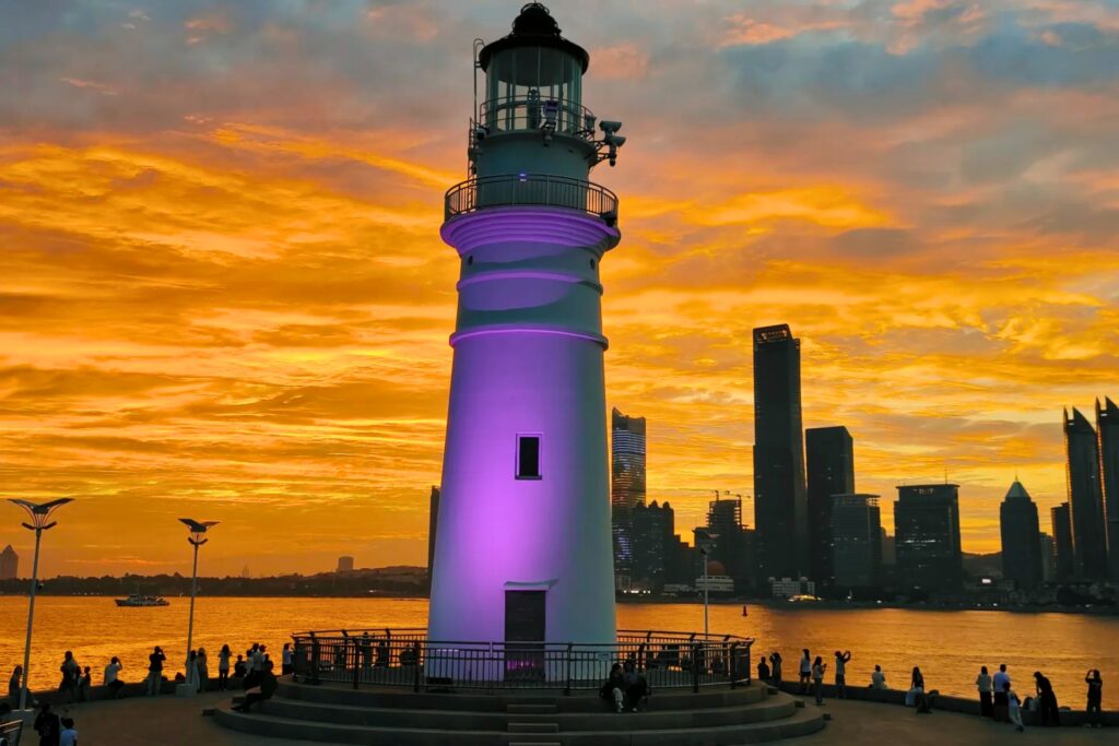 Phare blanc illuminé en violet sur la digue des Amoureux à Qingdao, avec un coucher de soleil orange spectaculaire et les gratte-ciel en arrière-plan