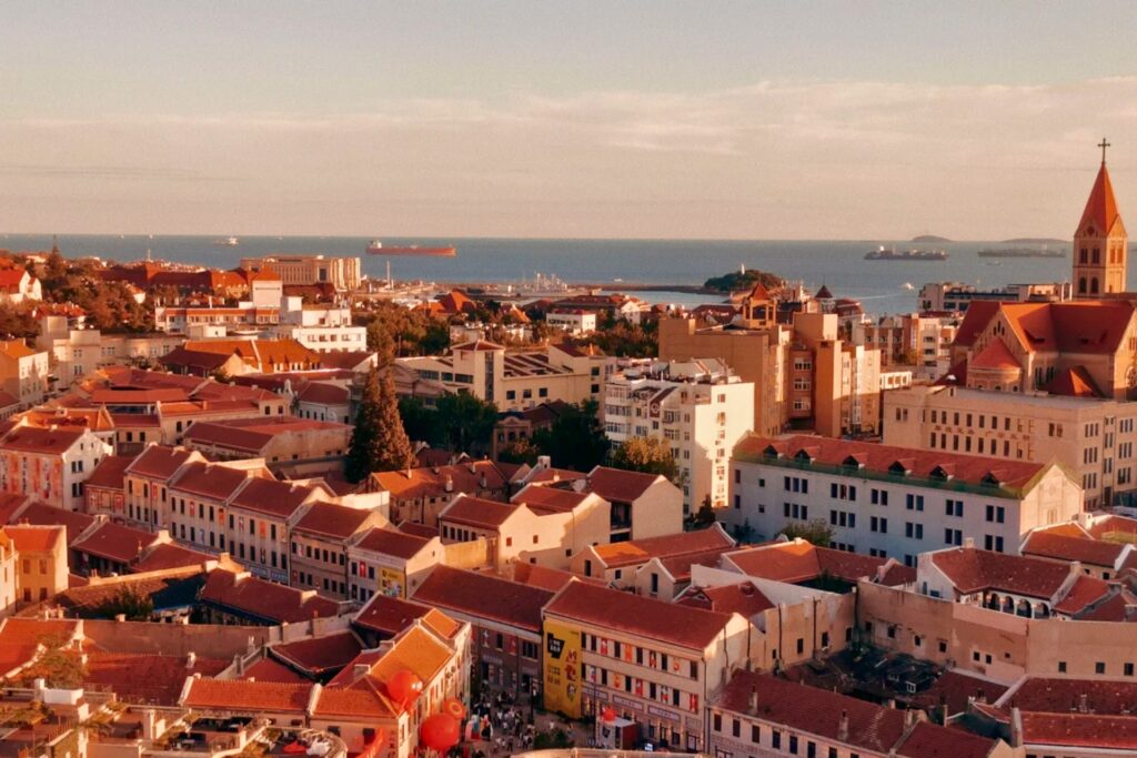 Vue aérienne du quartier historique autour de Qingdao Station, avec toits rouges, mer et cargos au loin sous une lumière dorée de fin de journée